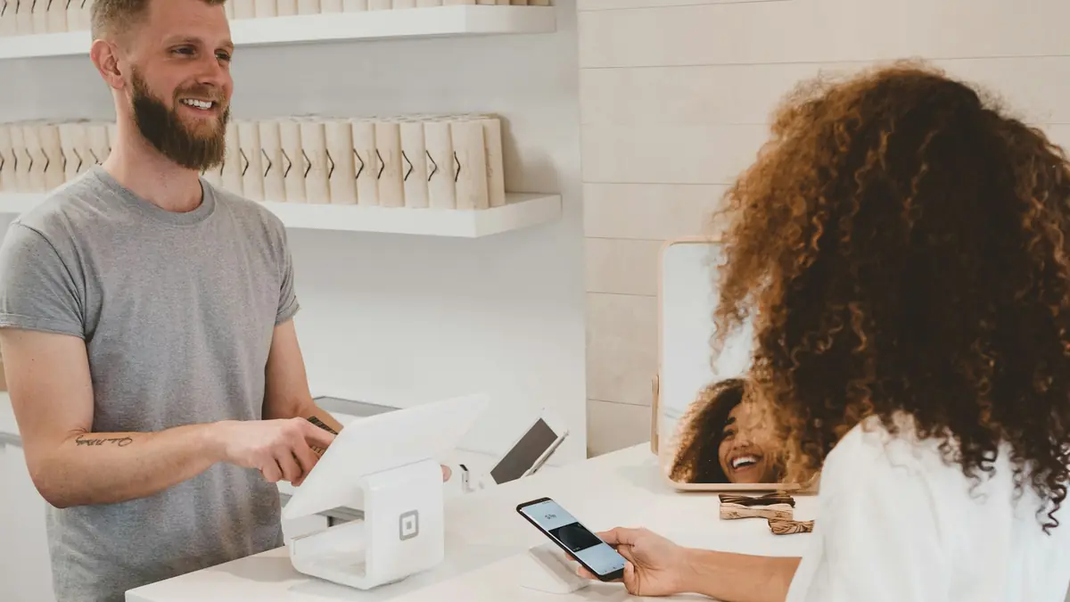 A shop owner smiles while a customer pays with a phone at the counter.