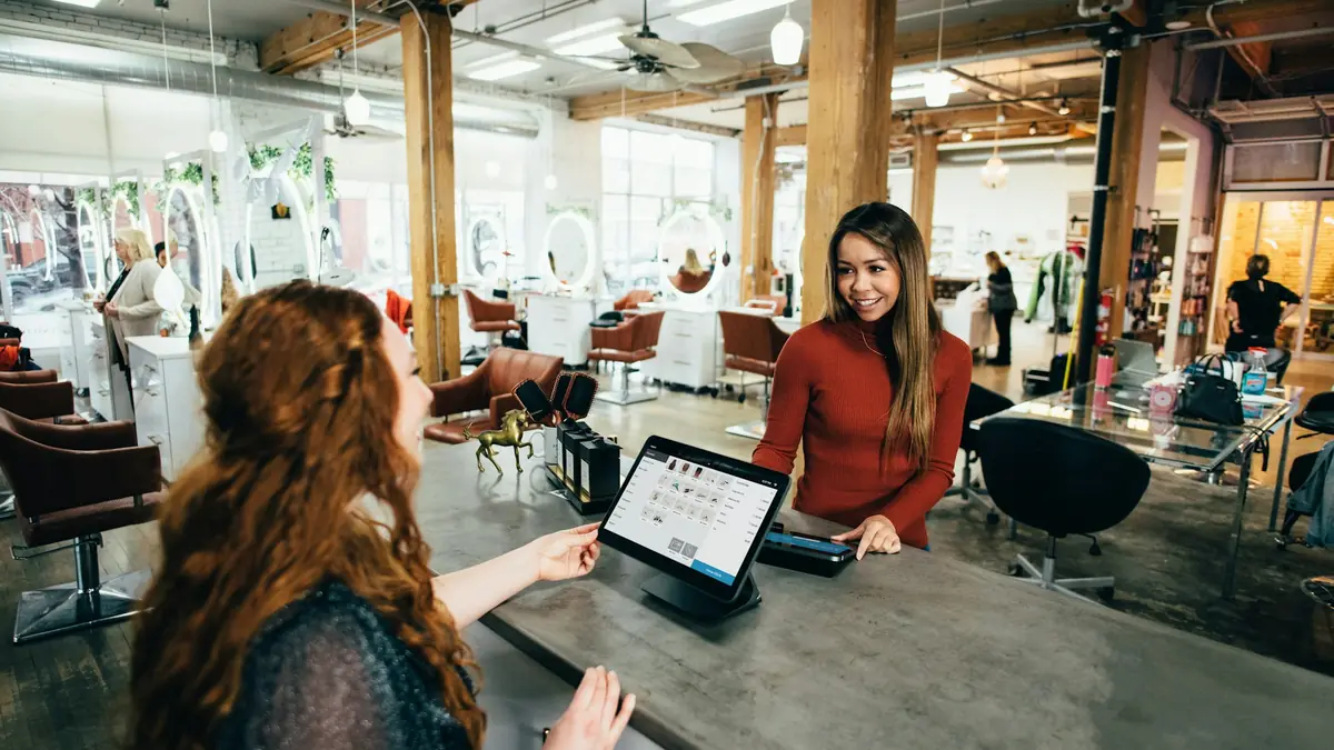 Two women talk across a front desk with a tablet in a bright studio.