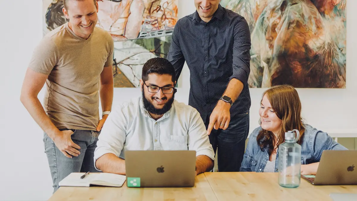 Four colleagues gather around a laptop on a shared table.
